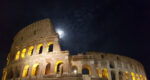 The Colosseum in Rome. The sky is dark and the moon is rising behind it