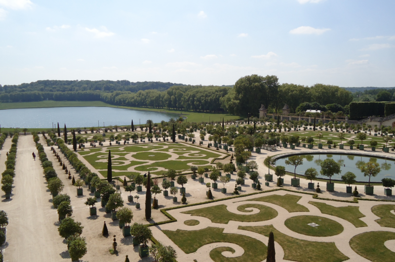Ornate gardens at the Palace of Versailles with hedges cut in circles and swirls in front of a pool of water